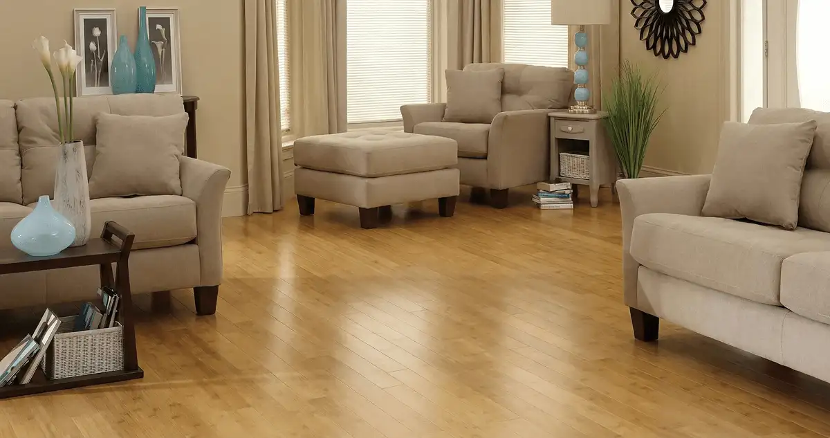 A living room with professionally installed bamboo flooring, beige sofas, a matching ottoman, a side table with a blue lamp, vases, framed pictures, and large windows with blinds and curtains in San Francisco, CA